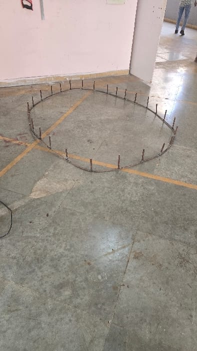 The image displays an indoor scene, likely a large room or hall, with a light pink wall on the left and a concrete or tiled floor covering the majority of the frame. The floor is marked with faded yellow lines, possibly indicating a court or designated area, and shows signs of wear and dirt.
A prominent object on the floor is a dark, thin, flexible material, possibly a metal strip or heavy wire, bent into an irregular oval or teardrop shape. Along its perimeter, numerous small, dark, rod-like pieces are attached vertically and spaced somewhat evenly. Another dark electrical cable or cord is visible, loosely coiled on the floor in the lower right corner.
In the upper left portion of the image, the lower body of a person is partially visible. They are wearing blue jeans and dark shoes, with a reflection on the polished floor surface.
No specific activity is clearly depicted, but the arrangement of the bent object with the vertical rods suggests a setup for an experiment, a boundary marker, a model, or an installation of some kind. The lighting appears to be typical for an indoor daytime setting.
A faint, partially obscured character, possibly 'a' or 'd', in a dark red or black color, is visible on the lower left pink wall. The overall setting, combined with the location context of Gangapur, India, could suggest a public building, perhaps a school, workshop, or community center.