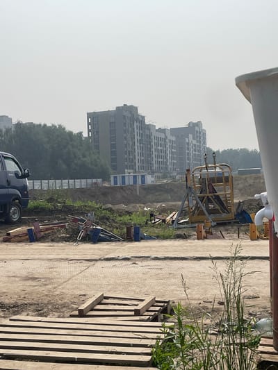 The image depicts a construction site in Koltsovo, Russia, characterized by a hazy sky and unfinished buildings in the background. In the foreground, a blue truck is parked next to a pile of construction materials, including wooden planks and metal components. A yellow metal structure, possibly part of a lifting device, stands prominently in the middle ground. Several portable toilet units are visible in the distance. The ground is a mixture of dirt and paved surfaces, with vegetation growing around the edges. The overall scene suggests ongoing construction activity in a developing area.
