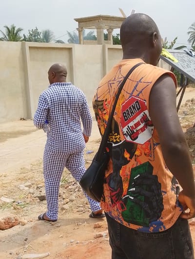 Two adult males are depicted from behind, outdoors in Agukwu Nri, Nigeria. In the immediate foreground, facing away from the camera and slightly to the right, is a bald male wearing an orange sleeveless t-shirt featuring a multi-colored graphic design including the text "BRAND PLEASE STOP COUNTERFEIT," skulls, flames, and a skeletal hand holding what appears to be green currency. He wears dark jeans and has a black sling bag draped over his left shoulder. Further into the scene, walking away from the camera on a reddish-brown dirt path, is another bald male dressed in a blue and white checkered long-sleeved shirt and matching trousers, with dark sandals. He carries a clear plastic water bottle in his left hand. The ground is uneven dirt with scattered small rocks and debris. In the background, a light-colored concrete wall spans horizontally. Behind this wall, an ornate, light-colored structure with columns, possibly a partially constructed gazebo or monument, is visible, with a faint wisp of smoke or dust near it. To the right, a metallic frame structure, possibly for solar panels, stands, with a light-colored piece of fabric draped over it. Palm trees and other green foliage are visible against an overcast sky in the distance.