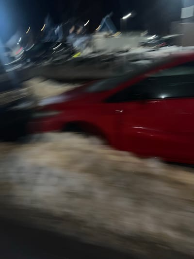 The image displays a blurry, low-light nighttime scene in New Haven, United States of America, characterized by significant motion blur. A red sedan, occupying the mid-ground, appears positioned against or partially within a substantial accumulation of dirty, partially melted snow or slush. The immediate foreground consists of a dark surface, likely asphalt, partially covered by the snow accumulation. In the highly blurred background, multiple vehicles are discernable, suggesting a commercial automotive environment such as a dealership or a large parking lot. Elongated streaks of artificial light sources, consistent with streetlights or building illumination, are visible across the distant background. No individuals are clearly identifiable within the frame. The scene depicts winter conditions. The precise street address or specific landmark within New Haven is not discernible from the image, but the setting is consistent with a commercial or industrial zone.