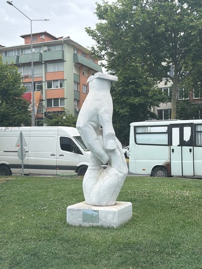The image captures a white marble sculpture depicting two clasped hands, positioned on a square base in a grassy area. Above the hands, a small rectangular object, possibly a book, is balanced. In the background, a multi-story building with orange and green facades and numerous windows is visible, alongside a street lamp. To the left, a white van is parked, and to the right, a white bus with black window frames is present. The overall scene appears to be an urban environment, likely a park or public space. This sculpture is located in the Süleymanpaşa district of Tekirdağ, Turkey, specifically in the vicinity of the Tekirdağ Museum of Archaeology.