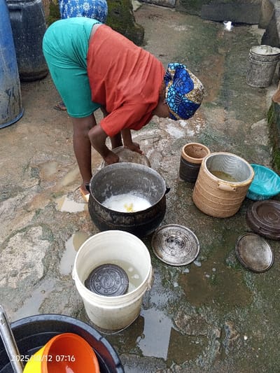 The image, captured in Kamantan, Nigeria, on 2025/10/12 at 07:16, displays an outdoor scene focused on an adult female engaged in a task. The woman, wearing a rust-orange short-sleeved top, teal-green shorts, and a blue and brown patterned headwrap, is bent forward with her hands positioned near a large, dark, roughly spherical pot. This pot contains a white foamy liquid with yellow solid material submerged within it.
The ground is a wet, uneven grey concrete or stone surface with numerous puddles and reflective areas. Surrounding the central pot are various containers and objects. To the lower left, a white plastic bucket holds water and a dark, circular lid with a raised pattern. Several other dark, circular lids, some with patterns, are resting directly on the wet ground. To the right of the central pot, a light brown cylindrical container with a textured surface and a metallic insert holds a yellowish liquid, and a smaller, dark brown cylindrical container stands nearby. A light blue plastic basin is also visible on the ground. In the background to the left, two blue plastic barrels are partially visible. Further in the background, a concrete structure with some green growth and another grey bucket are present. In the immediate foreground, a black plastic basin contains an orange cup and other indeterminate items.