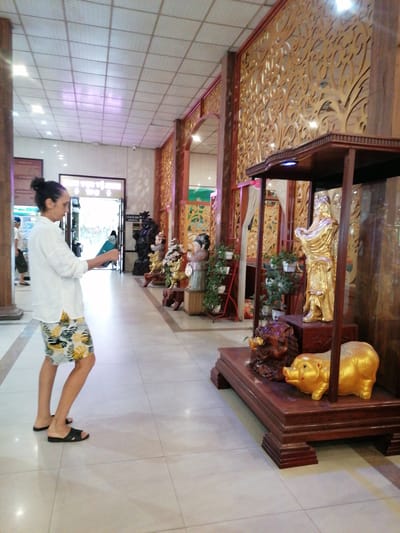 A person in a white shirt and patterned shorts stands in a building in Chu Văn An Ward, Vietnam. They are facing away from the camera, towards an open doorway at the end of a hallway. The hallway is lined with dark wood paneling with decorative cutouts on the right side, interspersed with statues and displays. A large display case with a gold statue and a pig is in the foreground on the right. The floor is tiled.