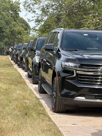 A line of black SUVs is parked on a textured surface next to a grassy area under a canopy of trees. The perspective is low, focusing on the front of the closest SUV, which has chrome accents on its grille and bumper. A man wearing a light-colored patterned shirt and dark pants stands in the background, looking down at something in his hands, positioned to the left of the parked vehicles. The environment appears to be outdoors, possibly a park or a roadside, with dry grass and lush green trees. The sky is visible through the branches, suggesting it is daytime. The scene is in the Town of Southampton, United States of America.