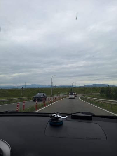 The image depicts a view from inside a vehicle driving on a highway. The road curves to the right, with a line of orange and white traffic cones marking the shoulder on the left. A gray SUV is visible on the road ahead, in the left lane. Further down the road, a white minivan and another vehicle are traveling in the right lane. The landscape on either side of the highway consists of green fields and trees, with a line of mountains in the distance under a cloudy sky. The foreground shows the dashboard of the car, with a decorative object on the right side. There is no discernible information within the image to suggest the location is specifically Samtredia, Georgia.