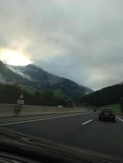 The image depicts a highway scene in the Austrian Alps, likely near Rothenwand, with overcast skies and dramatic cloud formations over misty mountains. A black SUV with the license plate "BK5-3233" is in the foreground, driving on the right side of the road. In the distance, other vehicles are visible on the highway, and a road sign indicating "300m" with a warning triangle is positioned to the left. The surrounding environment consists of steep, forested hillsides and a concrete barrier along the highway. The overall atmosphere is moody and somewhat dramatic, characteristic of mountain weather.