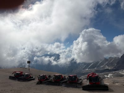 The image depicts a mountainous landscape with several red snow groomers parked on a gravelly surface. In the background, dramatic clouds hang over rugged peaks, with some patches of snow visible on the slopes. The sky is a mix of blue and white, with sunlight breaking through the clouds. A signpost with a dark display screen and two benches are visible to the left of the snow groomers. The scene appears to be an outdoor area, possibly a ski resort or a mountain pass, given the presence of the snow grooming vehicles. The location is identified as being in Wildermieming, Austria. There are no people visible in the image.
