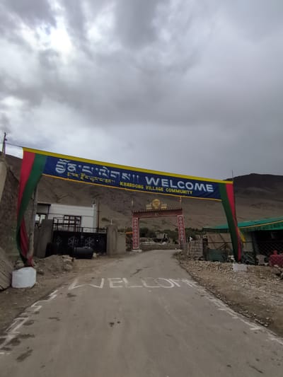The image depicts an entrance archway to Khardung Village, India, with a large banner displaying "WELCOME" in English and Tibetan script. The banner is suspended across a road that leads into the village. The road surface has "WELCOME" painted on it in white capital letters. The archway itself is adorned with a decorative frieze and Buddhist prayer flags. To the left of the road, there is a building with a white facade and a metal gate. To the right, a green awning covers a structure. The background shows a mountainous, arid landscape under a cloudy sky. There are no people visible in the image.