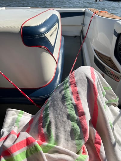 A first-person perspective image depicts the interior of a recreational powerboat on water in Coruche, Portugal, under bright daylight. In the foreground, the upper legs of an individual are partially visible, covered by a white terry cloth towel featuring prominent vertical stripes in neon green and coral pink. To the left, a white marine seat with dark blue sections and red piping is present, displaying a "NAUTIQUE" brand logo on its side. To the right, another section of the boat's white interior hull features dark brown textured marine flooring and a circular speaker grill. A red and white braided rope extends diagonally across the boat's floor from the upper left to the lower right of the frame. The background shows a body of water with a gently rippled surface, suggesting the boat is afloat on a river or lake. A blurred, dark shape is faintly visible on the water in the distant background.