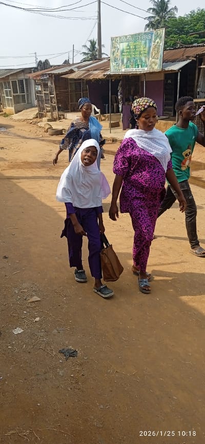 An image captured in Ifo, Nigeria, depicts a daytime scene on an unpaved, reddish-brown dirt road. Four individuals are present, walking from left to right across the frame.
In the foreground, a young person, likely a child, wears a white hijab over a purple long-sleeved top and trousers, along with dark-colored clogs bearing an "Adidas" logo. This individual carries a brown tote bag in their right hand and is looking towards the camera with an open mouth. Adjacent to them on the right is an adult female, wearing a vibrant purple and black patterned top and trousers, a white sheer headscarf with polka dots, and a multi-colored beaded headpiece. She wears dark sandals. To her right, an adult male wears a green t-shirt with a cartoon bear graphic and dark trousers, also with sandals. He holds a clear plastic item to his mouth, appearing to drink or blow. In the mid-background, another adult female walks, dressed in a blue and dark-patterned outfit with a matching head covering.
The background features a row of single-story commercial or residential structures constructed with a mix of corrugated metal and other materials, topped with corrugated metal roofs. One prominent green and white sign for "MAMA TEE BAR" is visible, listing services such as "WINE BEER COT Pepper Soup Fried Meat Poundo Local Gins Bitters Herb Pile, Opa Eyin, Ale etc." and two phone numbers. Utility poles with multiple overhead wires traverse the scene above the buildings. Sparse vegetation, including palm trees, is visible behind the structures. The sky is clear. Scattered debris, including small pieces of litter and dried plant matter, is present on the dirt road. The image is timestamped "2026/1/25 10:18."