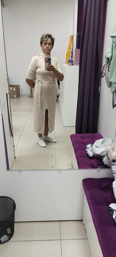 In a fitting room, a woman with short, styled hair stands in front of a mirror, taking a selfie with her phone. She is wearing a light-colored, knee-length dress with a tie at the waist and a high neckline. The dress has short sleeves and a slit in the front. She is also wearing white socks and white flat shoes.
The fitting room appears to be in a store, with white walls and a tiled floor. A purple curtain hangs on the right side of the room, and a purple cushioned bench is visible, with some clothing and sunglasses placed on it. To the left of the woman, a cardboard box is stacked on the floor, and a black trash can is also present. In the background, on a white shelf, there is a small potted plant with purple flowers and some other items, including what looks like a measuring tape. The lighting suggests it is daytime. The woman's expression is neutral. There is no visible text in the image.