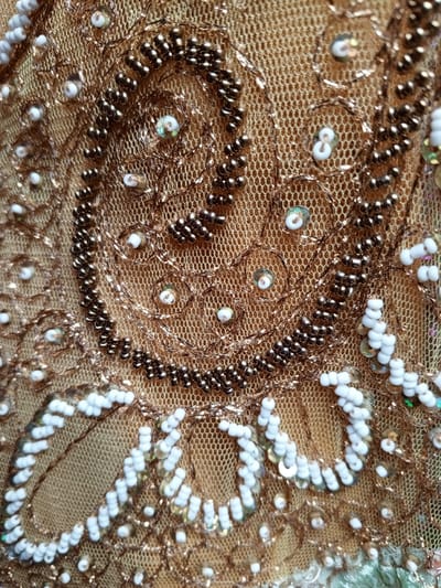 An ornate textile features intricate beadwork and sequins on a gold-toned net fabric. Dark brown beads form swirling patterns, complemented by smaller white beads and iridescent sequins scattered throughout. Fine metallic gold thread outlines the designs, enhancing the textured, luxurious appearance. The combination of embellishments creates a dense, decorative surface.