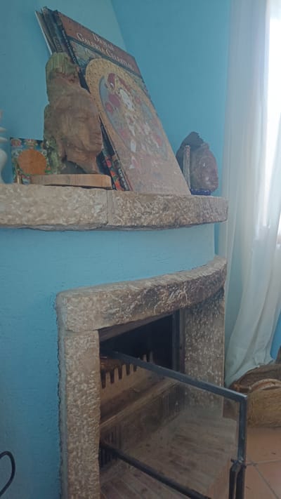 The image shows a stone fireplace in a room with blue walls. On the mantelpiece, there are books titled "DIOSAS GALERIA CELESTIAL" stacked behind a small stone sculpture of a head and a small painting. The fireplace opening contains a metal grate. A sheer white curtain is visible to the right of the fireplace. The location is Jérica, Spain.