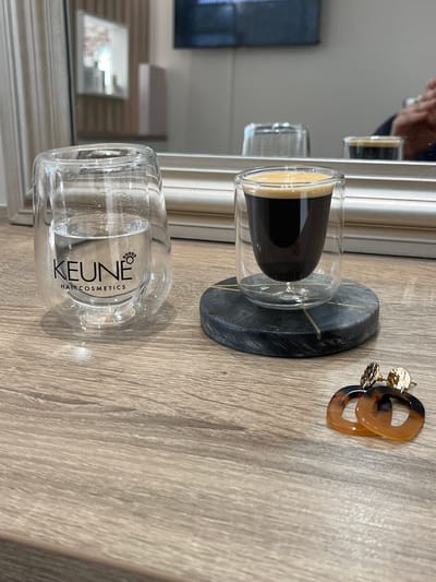 The image shows a close-up of a light-colored wooden surface. On the surface, there is a clear double-walled glass containing a dark liquid with a lighter-colored foam on top, possibly espresso. The glass is resting on a small, dark gray coaster with gold-colored veining. Next to the coaster are a pair of hoop earrings with a tortoise-shell pattern and gold-colored studs. In the background, there is a mirror reflecting the scene and revealing parts of a room with a TV screen on the wall and a framed picture. There is no specific information in the image to confirm the location in the city of Montrond-les-Bains, France.