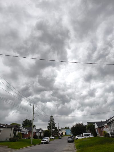 This is a photograph of a residential street in Hawkesbury, Canada, under a heavily overcast sky. The scene depicts a suburban neighborhood with houses on either side of a paved road. The houses are predominantly single-family dwellings with manicured lawns and trees. Several vehicles are parked on the street and driveways, including a white SUV, a dark sedan, and a white van. Utility poles with numerous wires crisscross the frame, indicating the presence of overhead power and communication lines.
The weather appears to be inclement, with thick, dark clouds dominating the sky, suggesting an impending storm or rain. The lighting is diffused, creating a somber mood. The time of day is likely daytime, but the overcast conditions prevent strong shadows. The overall impression is one of a quiet, ordinary suburban day, perhaps interrupted by changing weather. There is no visible text in the image.
