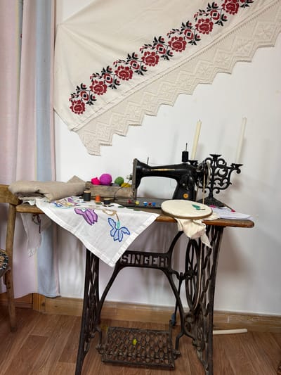 The image shows a close-up view of a sewing machine and surrounding crafting materials on a wooden table. The sewing machine is black and appears to be an antique Singer model. Various spools of thread, skeins of colorful yarn, and buttons are scattered on the table. A piece of white fabric with embroidered blue flowers is draped over the edge of the table, and another piece of fabric, folded, is on a wooden chair to the left. Above the sewing machine, a white textile with red floral embroidery and lace trim is hung on the wall. Two white candles in a decorative black candelabra stand behind the sewing machine. The scene is set against a plain white wall and a wooden floor. The overall impression is of a crafting or sewing area, possibly a workspace or a display in a home or small shop. The specific location in Ust'-Kabyrza, Russia is not determinable from the image itself.