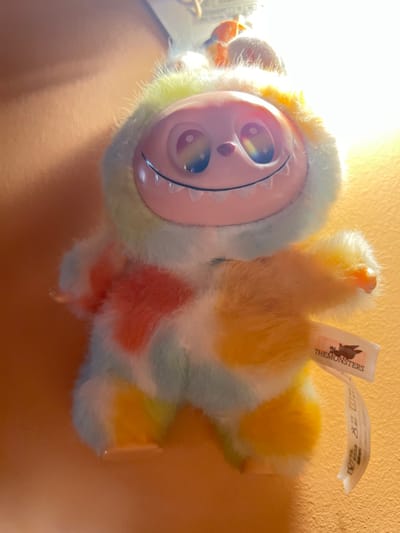 The scene depicts a multi-colored plush toy, approximately 15-20 cm in height, suspended against a plain, warm-toned background within the Gerakas Municipal Unit, Greece. The toy features a fluffy, patchwork fur body in light blue, white, yellow, orange, and green hues. Its face is light pink plastic, exhibiting large, dark, reflective eyes with rainbow-like reflections, and a wide, upturned smile revealing white teeth. Small, translucent plastic hands and feet are visible. A white fabric tag protrudes from the toy's right side, bearing the text 'THE MONSTERS' and an associated logo. Another partially torn tag hangs below it. A bright, localized light source illuminates the toy from above, casting a glare across its upper face. No other subjects or actions are present.
