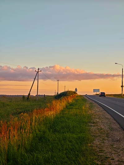 This image depicts a rural road scene in the vicinity of Kuronovo, Russia, during a sunset. The sky is a gradient of pale blue to soft orange, with wispy, pink-tinged clouds. The foreground is dominated by a lush green field with tall grass, some of which is catching the golden light of the setting sun. To the right of the field, a paved road curves gently into the distance. A dark-colored car is visible on the road, heading away from the viewer. On the left side of the road, a power line pole with wires extends across the frame. Further in the distance, another power line pole and what appears to be a distant forest are visible. A large, blank billboard is positioned on the right side of the road, near a street lamp. The overall atmosphere is serene and tranquil, characteristic of a rural landscape at dusk.