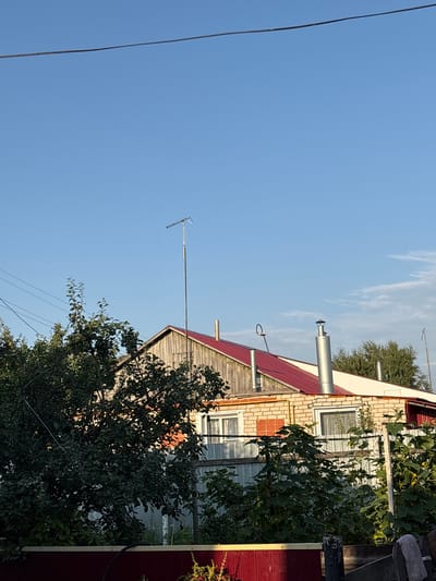 The image shows a rustic, rural scene under a clear blue sky with a few wispy clouds. A two-story house with a red metal roof is visible, constructed with light-colored bricks on the lower level and weathered wooden planks on the upper gable. Two chimneys, one with a conical cap, protrude from the roof. A television antenna stands tall on a pole near the peak of the roof. Lush green trees and foliage partially obscure the house, with dense bushes and plants in the foreground. A red fence runs along the bottom of the frame. The scene appears to be set in a village or countryside location, consistent with the provided location of Leyptsig, Russia. There are no people or animals visible in the image.