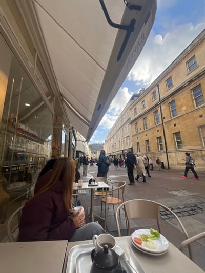 This image depicts an outdoor cafe scene on a pedestrianized street in the city of Bath, United Kingdom, specifically likely on Cheap Street. The perspective is from a low-angle at an outdoor table looking along the street.
In the immediate foreground, a metal tray holds a black teapot and a white ceramic mug containing a dark liquid, presumably tea. To the right of the tray, a white plate with remnants of a small salad (lettuce, a tomato slice, and a cucumber slice) rests on stacked plates. The table is light-colored.
Seated at an adjacent cafe table on the left, a woman with long, straight brown hair, wearing a dark maroon padded jacket and grey trousers, holds a white paper cup in her right hand. A gold ring is visible on her right ring finger. Her gaze is directed down the street. Beyond her, another cafe table is visible, with a person partially obscured and a small black moka pot on the table.
Overhead, a beige awning extends from the building, displaying the reversed word "BAKERY" in black lettering. The building on the left features large glass display windows reflecting the opposite side of the street and interior lights. Architectural details consistent with Georgian Bath stone buildings are visible.
The midground reveals a busy, brick-paved pedestrian street with multiple individuals walking in various directions. On the right, a long block of multi-story Georgian buildings, constructed from honey-colored Bath stone, features numerous symmetrical sash windows. Pedestrians populate the street, including a person in a dark jacket and another in a light-colored coat conversing, and a person in a grey jacket and bright pink footwear walking away from the camera on the far right.
In the background, the street recedes, lined by more similar Bath stone buildings. A distant, tree-covered hill or elevated terrain is visible under a partially cloudy blue sky.