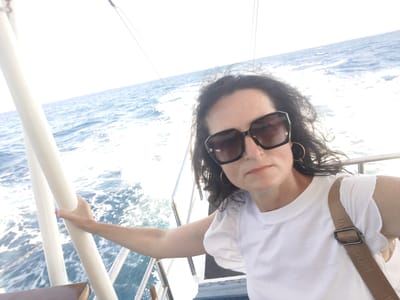 A woman with dark, wavy hair and large, dark-rimmed sunglasses is taking a selfie on a boat. She is wearing a white t-shirt with ruffled sleeves and a brown crossbody bag. She is holding onto a white railing. The ocean is visible in the background, with waves and white foam. The sky is overcast and bright. The woman appears to be in her 40s or 50s. The scene is likely taking place on the water near Kastri, Greece.