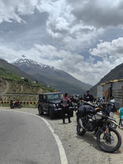The image captures a group of motorcyclists and their bikes parked on the side of a road with a dramatic mountain backdrop. In the foreground, a black Mahindra Thar SUV is parked. Several motorcycles are lined up, some with luggage attached, suggesting a road trip. People are visible around the vehicles, some standing and others possibly seated in a small roadside structure.
The scene is set in a mountainous region, with towering peaks, some capped with snow, dominating the background. Green slopes and rocky outcrops form the terrain leading up to the mountains. The sky is partly cloudy, with a mix of white and grey clouds, suggesting a daytime scene. The road appears to be paved with a white line marking the edge.
Visible text includes a license plate on one of the motorcycles which reads "SK-01PD-9997". Another motorcycle has "HIMALAYAN" written on it. Text on the jackets of some individuals is also visible but difficult to decipher clearly. The overall mood appears to be one of adventure and camaraderie, common among motorcycle touring groups. The location, with its rugged mountains and roadside stops, is indicative of a scenic route, likely in a region known for its challenging terrain and natural beauty, consistent with the context of India.