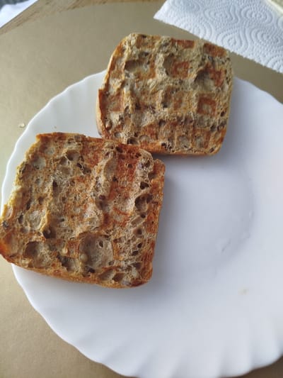 Two slices of toasted multi-grain bread are on a white, scalloped-edge plate. The bread is a light brown color with visible grains and seeds. The plate is placed on a beige, textured surface. To the right of the plate, a portion of a white paper towel is visible. This image was taken indoors, likely in a kitchen or dining area. There is no indication of the specific location within Offenbach am Main, Germany.