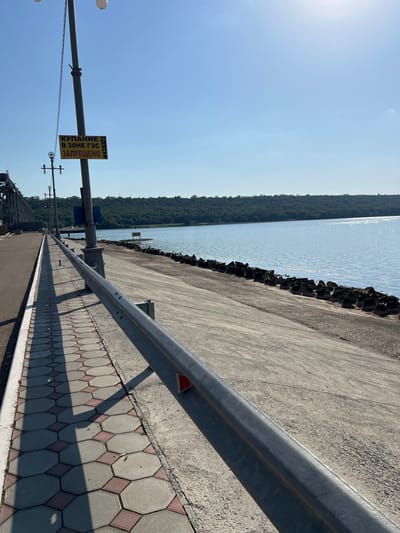 The image depicts a waterfront scene, possibly along a river or reservoir, with a clear blue sky. On the left, there is a paved walkway with a distinctive hexagonal tile pattern, bordered by a concrete embankment. A metal guardrail runs along the edge of the walkway. In the background, a large industrial structure, likely a bridge or dam, is visible. The water extends to the horizon, where a tree-covered shoreline forms a natural boundary. The sun is shining, casting long shadows from the guardrail and lampposts. A yellow sign on a lamppost reads "КУПАНИЕ В ЗОНЕ ГЭС ЗАПРЕЩЕНО", which translates to "SWIMMING IN THE HPP ZONE IS PROHIBITED". The location is identified as Korzhevo, Moldova.