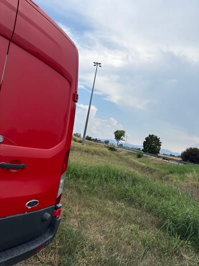A red Ford Transit van is parked on a grassy shoulder beside a highway in Fontevivo, Italy. The van is viewed from the rear passenger side, with the large red rear door and a portion of the side visible. The van has a black bumper with a visible Ford logo. A tall light pole stands in the background, beyond a sloped grassy embankment. The highway runs diagonally across the frame, with a metal guardrail separating it from the embankment. Several trees are scattered along the highway, and mountains are visible in the distance under a partly cloudy sky. The overall scene depicts a roadside environment in a rural or semi-rural area.