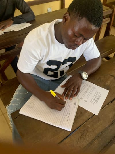 A young dark-skinned male is seated at a worn wooden desk, writing in a lined notebook with a yellow pen in his right hand. He is wearing a white t-shirt with a black "23" printed on the front, light blue denim jeans with silver embellishments on the left thigh, and a silver-colored wristwatch on his left wrist. To the right of his notebook, a white sheet of paper with visible text and a possible university logo is present. His gaze is directed downwards at his writing. In the background, another dark-skinned individual, partially visible, sits at an adjacent wooden desk, engaged with papers. The setting is an indoor room, likely a classroom or examination hall, characterized by light-colored walls and additional worn wooden desks. A white electrical outlet is visible on the wall. The scene is located in Abobo, Nigeria.