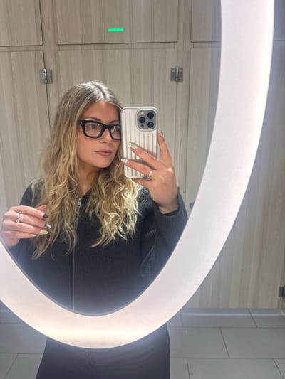 A blonde woman with glasses takes a selfie in a mirror with a circular light. She is wearing a dark gray, zip-up cardigan. Her phone has a beige, ridged case. She has rings on multiple fingers and decorative nail art. The background shows light-colored, wood-grain patterned cabinets with chrome hardware and white tiled walls. A rectangular green light is visible on the left cabinet door.