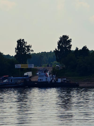 The image shows a wide, calm body of water in the foreground, with ripples reflecting the light. In the midground, a ferry or barge is docked at a shoreline with a sandy or muddy bank. The vessel appears to be a multi-decked structure, with a white cabin on the upper level. There are several vehicles visible on the ferry, including a white car and a red truck. Adjacent to the ferry, on the shore, there are buildings, including a small structure with a blue roof and windows. A large white trailer is also parked nearby. Behind the shoreline, a dense forest of dark green trees creates a backdrop, with two prominent trees with thin branches standing out against the lighter sky. A billboard with Russian text is visible amongst the trees. The sky is overcast and light-colored. The scene is likely a port or a ferry crossing in Erkalovo, Russia, given the presence of Russian text on the billboard and the general landscape.