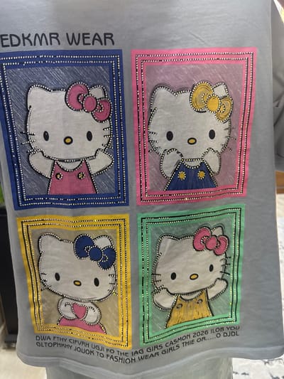 This is a close-up, eye-level shot of a light grey t-shirt featuring a repeating pattern of four illustrated Hello Kitty characters within decorative frames. The text "EDKMR WEAR" is printed in black uppercase letters above the illustrations. Each Hello Kitty is adorned with rhinestones along its outline and within its facial features, as well as around the frames. The frames are bordered by rhinestones and filled with solid colors: blue, pink, yellow, and green. The first Hello Kitty, in the top left blue frame, wears a pink dress. The second, in the top right pink frame, wears a blue dress with yellow accents. The third, in the bottom left yellow frame, wears a pink dress. The fourth, in the bottom right green frame, wears a yellow dress with green accents. Below the illustrations, there is a line of text which appears to be a brand name or slogan, partially obscured and difficult to fully decipher due to the fabric's texture and the angle. The t-shirt is likely an item of clothing, and the image does not provide any information about its location in Shofirkon, Uzbekistan, or any surrounding environment.