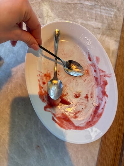 The image depicts a white, oval-shaped plate located in Mysiadło, Poland, showing remnants of a consumed dessert. The plate's surface is extensively smeared with a bright red, viscous, fruit-based residue, consistent with strawberries or berries, along with scattered white crystalline powder, likely sugar, and small white solid fragments. Two sliced strawberry pieces are visible on the left side of the plate. Two metallic, silver-colored spoons are present. One spoon lies horizontally on the plate, partially submerged in the red residue, its handle oriented upwards. The second spoon is held by a fair-skinned human hand, positioned in the upper left quadrant of the frame. The hand, with visible fingernails coated in clear polish, grips the handle of the spoon between the thumb and forefinger. This spoon is angled diagonally, with its bowl resting on the first spoon. A minor superficial scratch is visible on the dorsal surface of the index finger of the hand. The plate rests on a light-colored, veined stone surface, possibly marble or granite. A vertical section of a light-brown wooden surface is visible along the right edge of the image. The scene is illuminated, casting a shadow from the hand and spoon onto the stone surface.