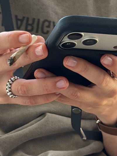 The media file shows a close-up of a person's hands holding a smartphone and a rolled cigarette. The person is wearing rings on their fingers, including a metallic ring with a unique, textured design on one finger and a simpler band on another. The smartphone, partially visible, appears to be in a dark-colored case and has multiple camera lenses. The rolled cigarette is held between the thumb and forefinger of one hand. The background consists of textured fabric, possibly clothing, in a neutral color. The lighting suggests an indoor setting. There is no discernible text visible in the image. The overall impression is casual and intimate, focusing on the tactile interaction between the hands and the objects they hold. The image is cropped closely to the hands and the objects, creating a sense of focus on these elements. The angle of the shot is slightly tilted, adding a dynamic feel to the composition.