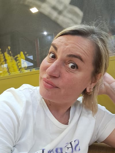 A woman with blonde hair and fair skin is looking directly at the camera. She is wearing a white t-shirt. The background appears to be a window with a yellow frame and some reflection of yellow objects outside. The location in Bel'kovo, Russia, is not evident in the image.
