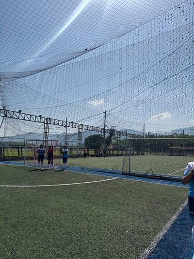This image captures an outdoor artificial turf sports complex in Pitalito, Colombia, under bright daylight conditions. The playing area features green artificial turf with white demarcation lines and a blue perimeter track. An extensive black protective netting system, supported by metal trusses and vertical poles, covers the entire field overhead; this netting exhibits visible accumulated debris.

In the mid-ground on the left side, three individuals are positioned near a goal frame with white netting. Two individuals wear light-colored sports jerseys (one blue, one white), while the central figure wears a red jersey and has their right arm raised. To their right, another goal structure with white netting is visible further back on the field. Approximately five additional individuals are present in the background, some standing and others seated near a dark fence that borders a natural landscape of trees and distant mountains. A partial view of a person in a light blue and white jersey is visible in the extreme right foreground, holding an indeterminate object. The sky is clear blue with sparse white clouds, and a diagonal light streak is visible across the upper left sky quadrant.