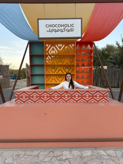 The image captures a female subject positioned behind a reddish-brown display counter within an outdoor setting in Hatta, United Arab Emirates. The individual has long dark hair and wears a white hooded top, with her hands resting on the counter's upper edge as she looks directly at the viewer. The counter's front features a decorative band with a red and white geometric zigzag pattern. Behind the subject, a multi-tiered display unit is visible, consisting of teal shelves on the left and larger sections in yellow and orange, both adorned with intricate geometric cut-out patterns. Overhead, a white sign with "CHOCOHOLIC" in English and Arabic text is suspended, accompanied by draped fabric in light blue, tan, and reddish-orange hues. The background shows sparse desert vegetation, including visible palm trees, rustic fencing elements, and a clear, light blue sky. The ground in the immediate foreground is a patterned stone pavement, transitioning to a lighter, possibly gravel or dirt, surface further back.