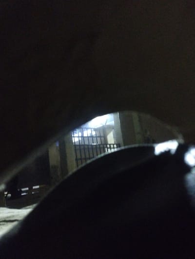 The scene, located in Kubare, Nigeria, is captured at night, characterized by low light conditions with a bright artificial light source in the background. The foreground features a dark, curved object, likely an internal component of a vehicle, which partially frames the central view. Through this opening, a metal gate with vertical bars is visible. Behind the gate, structural pillars of a building are present, beyond which the bright light source illuminates an area. To the left of the gate, two distinct, parallel white horizontal markings are discernible on a dark, level surface. No individuals are depicted, and no specific actions or interactions are occurring. The visible environment includes paved ground.