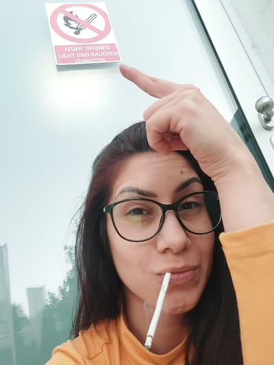 In Altdorf, Germany, a woman with dark hair and glasses holds a cigarette to her lips, with smoke rising from it. She is wearing an orange shirt and her right hand is raised, pointing at a sign that reads "FEUER, OFFENES LICHT UND RAUCHEN VERBOTEN," which translates to "FIRE, OPEN LIGHT AND SMOKING PROHIBITED." The sign is affixed to a glass surface, likely a window or door, which reflects the surrounding environment, including trees and a building. A metallic door handle is visible to the right of the frame. The overall scene suggests a moment captured in a public or semi-public space where smoking is prohibited.