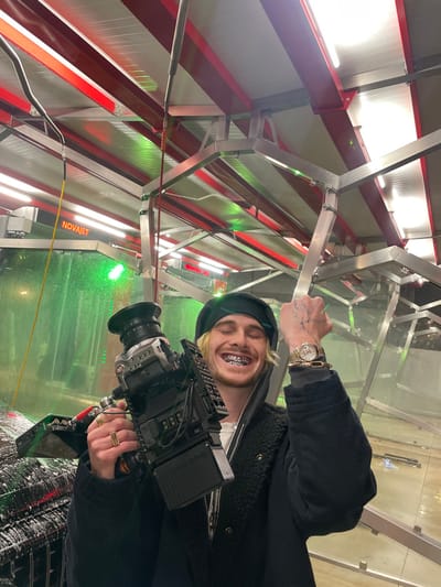A person wearing a beret, jacket, and diamond grill stands inside a car wash holding a large camera. The person has a gold watch and hand tattoos. The car wash structure is red, white, and silver with lights overhead. The wall features green illuminated water jets. The camera is aimed at the spraying water.
