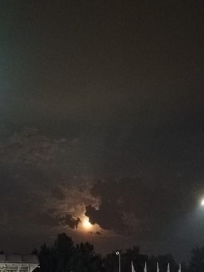 The image captures a nocturnal scene of the sky above Bytom, Poland, with the moon partially obscured by dark, textured clouds. The moon emits a warm, orange glow, creating a dramatic contrast with the surrounding darkness. Below the sky, silhouetted against the faint light, are the tops of trees and what appears to be a structure with a roofline and supporting beams on the left side of the frame. On the right side, a bright, artificial light source is visible, likely a lamp or stadium light, casting its illumination upwards. In the lower portion of the image, several white flags on poles are arranged in a line, suggesting an outdoor event or location. The overall atmosphere is moody and somewhat mysterious, characteristic of a night sky with cloud cover.