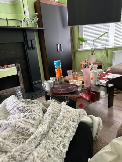 The scene is an indoor living space located in Dorchester, United Kingdom. The room features light green walls and light wood laminate flooring. On the left, a black fireplace mantelpiece is present, adorned with a dark candle holder containing a green candle and three silver and blue decorative trophies or figurines. To the right of the fireplace, a tall, dark brown and black wardrobe with two vertical silver handles stands against the wall. A wall-mounted television is partially visible at the top center of the frame. A window with white horizontal Venetian blinds is visible on the right, with a white radiator positioned beneath it. A small light-colored side table near the window holds a potted plant with broad green leaves.
In the mid-ground, a cluttered glass-top coffee table with black legs is prominent. Resting on the table are: a maroon oval plate with food remnants and a silver fork; a white ceramic mug; an orange Pringles can; a salt shaker and a pepper shaker; a pink water bottle with a black lid; multiple clear plastic cups, one with a McDonald's logo; a small bottle containing liquid, possibly vape juice; a wooden circular board; an open red box, partially obscured but showing "Heartland" and "10"; and various crumpled red and white food wrappers.
In the immediate foreground, a person's legs, dressed in dark trousers, are visible, partially covered by a light grey, fluffy textured blanket, indicating an individual is seated on a sofa or armchair.