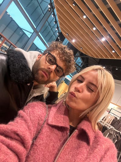 A close-up selfie captures two individuals inside a modern retail store, identified as likely within the Cap3000 shopping center in Saint-Laurent-du-Var, France, based on discernible "CABINES D'ESSAI" (fitting rooms) signage and contemporary architectural features.
On the left, a male subject with curly brown hair, a beard, and black-framed glasses wears a dark jacket with a black faux fur collar over a grey hooded sweatshirt. He maintains a direct gaze at the camera.
On the right, a female subject with shoulder-length blonde hair wears a textured, fuzzy pink coat with a silver zipper and a visible silver chain strap, along with a gold-colored earring in her left ear. She is depicted winking her right eye and puckering her lips towards the camera.
The background reveals sections of the store's interior, including clothing racks displaying garments, a high ceiling with both translucent glass panels and wooden slats containing recessed spotlights, indicative of a commercial environment.
