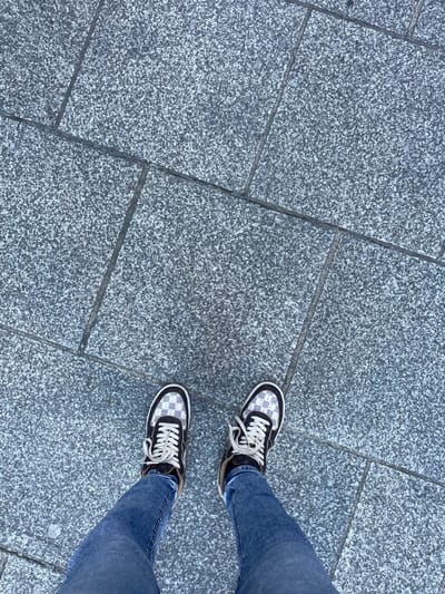 An overhead shot depicts a person's lower legs and feet standing on a paved surface in Zarautz, Spain. The individual is wearing blue denim jeans and dark-toned sneakers featuring a prominent checkerboard pattern in light grey and white, accented with dark trim and off-white laces. The person's feet are positioned slightly apart, indicating a stationary stance. The ground consists of irregularly shaped, light grey granite paving tiles with visible dark grout lines, exhibiting a finely speckled texture. The ambient lighting appears even, suggesting diffused sunlight or an indoor setting.