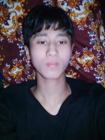 A head-and-shoulders portrait features a young individual, likely male, with fair skin, short dark hair, and dark eyes, looking directly into the camera with slightly pursed lips. The individual wears a plain black V-neck long-sleeved shirt. The subject is positioned against a patterned textile, possibly a pillow or blanket, which displays repeating gold-yellow scrollwork and orange-red floral designs with green leaves set against a dark reddish-brown background. Some blue floral elements are also present in the textile pattern. The specific location within the city of Bagat, Uzbekistan, cannot be determined from the visual evidence in the image.