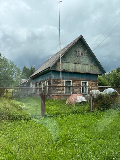 A dilapidated wooden house with peeling green and brown paint stands under a cloudy sky in Шапечынскі сельскі Савет, Belarus. The house has a pitched roof covered in weathered wooden shingles. There are several windows on the facade, some with glass and others with broken panes. A tall wooden pole, likely for an antenna, is planted near the house. A chain-link fence runs across the foreground, separating the viewer from the property. Overgrown green grass and weeds cover the ground. To the right of the house, several objects are covered with white and red tarps, suggesting they are stored outdoors. Trees and foliage surround the house, and another smaller building with a grey roof is visible in the background to the left. The overall scene conveys a sense of disrepair and rural abandonment.