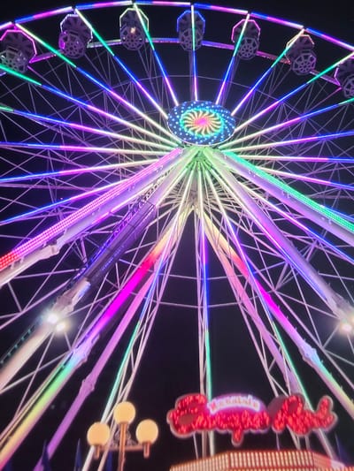 A large Ferris wheel, brightly illuminated with a dynamic array of LED lights, dominates the night scene. The Ferris wheel's central hub features a vibrant, swirling psychedelic pattern of blue, green, yellow, pink, and red lights. Spokes radiate outwards from the hub, glowing in various shades of green, blue, purple, and pink. Many of the structural beams of the wheel are also outlined or covered with these colorful lights.

Numerous passenger gondolas or cabins are visible along the perimeter of the wheel, each illuminated with subtle light. The dark background emphasizes the vividness of the lights, indicating it is nighttime. To the right side of the Ferris wheel, partially visible against the dark sky, are elements of another structure or ride. This includes large, red illuminated shapes that appear to be stylized letters or decorative elements, though they are partially obscured and difficult to decipher clearly. Below these red elements, several round, warm yellow lights on poles are also visible.

The overall impression is that of a lively amusement park or fairground. The diverse colors and intense lighting create a festive and vibrant atmosphere. No specific text is clearly legible on the red illuminated shapes.