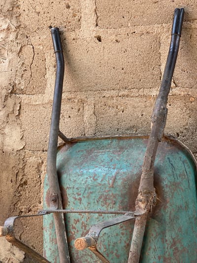 The image shows the handles and top of a weathered, teal-colored wheelbarrow leaning against a textured, tan-colored wall. The wall appears to be made of concrete or stucco with visible cracks and an uneven surface. The wheelbarrow exhibits signs of rust and wear, particularly on its metal frame and handles. The handles are dark gray, with black plastic grips at the ends, and show some damage. A horizontal metal bar is visible across the top of the wheelbarrow. The scene appears to be an outdoor setting, possibly a yard or an alley, in a location consistent with Daniel, Nigeria, given the architectural style of the wall and the likely use of such a tool in that environment. No people or specific actions are depicted.