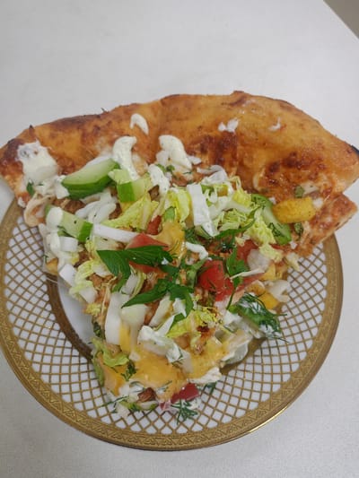 A slice of pizza is presented on a decorative plate. The pizza crust is golden brown with some charred edges. It is topped with a generous portion of salad, consisting of shredded lettuce, chopped tomatoes, cucumbers, and onions. A creamy white dressing is drizzled over the salad and pizza. Some fresh herbs, likely dill and parsley, are scattered throughout the toppings. The plate has a white base with an intricate gold geometric pattern around the rim. The background is a plain white surface, suggesting an indoor setting. No people are visible in the image. The location in the city of Avtury, Russia, is not discernible from the provided image.